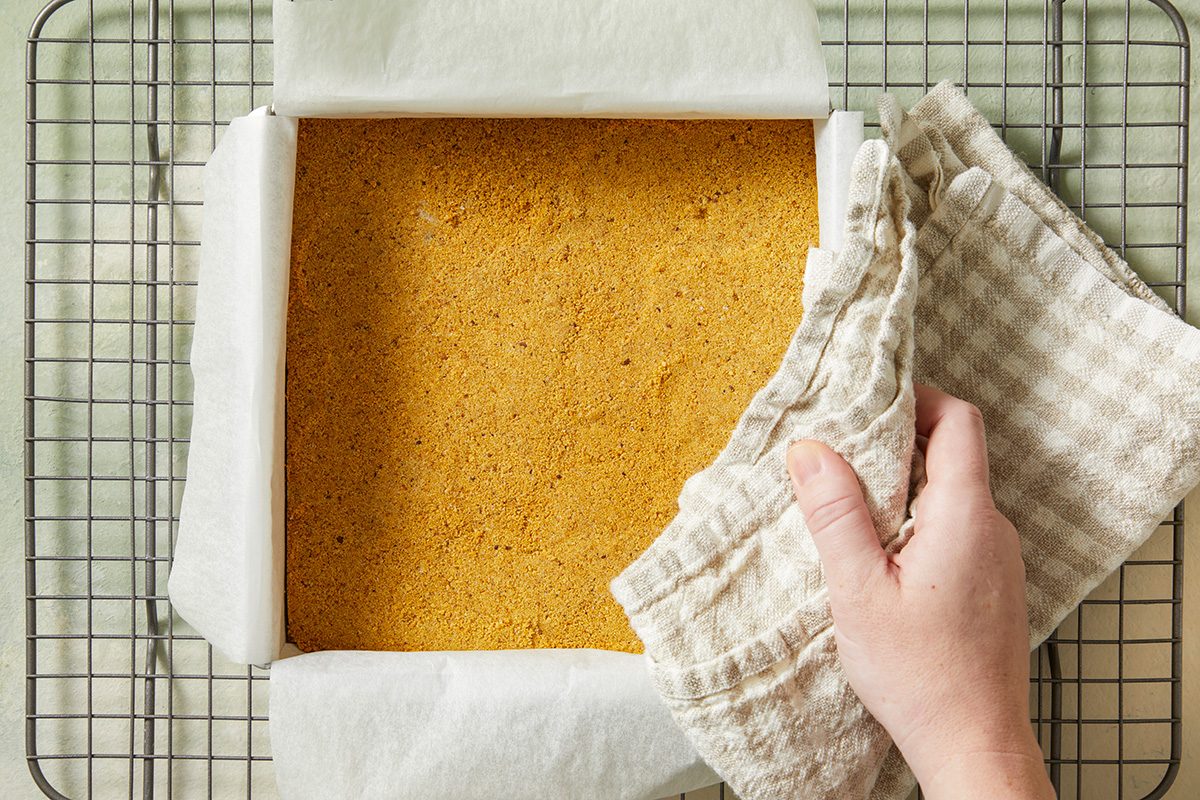 A hand using a checkered cloth lifts a square pan lined with parchment paper, filled with a golden-brown baked cake, resting on a wire cooling rack.