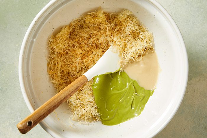 A white bowl contains shredded phyllo dough, beige liquid, and green paste, with a white spatula resting inside, ready to mix the ingredients.