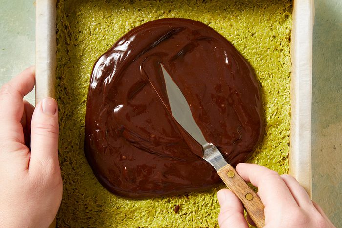 A person spreads melted chocolate over a green cake base in a rectangular baking pan using a spatula.