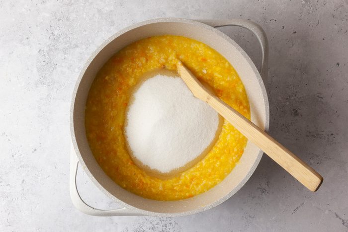overhead shot of a pot filled with a yellowish mixture that appears to be a blend of ingredients such as squash; in the center of the mixture, there is a mound of white sugar; a wooden spatula rests against the edge of the pot, the background features a subtle gray surface