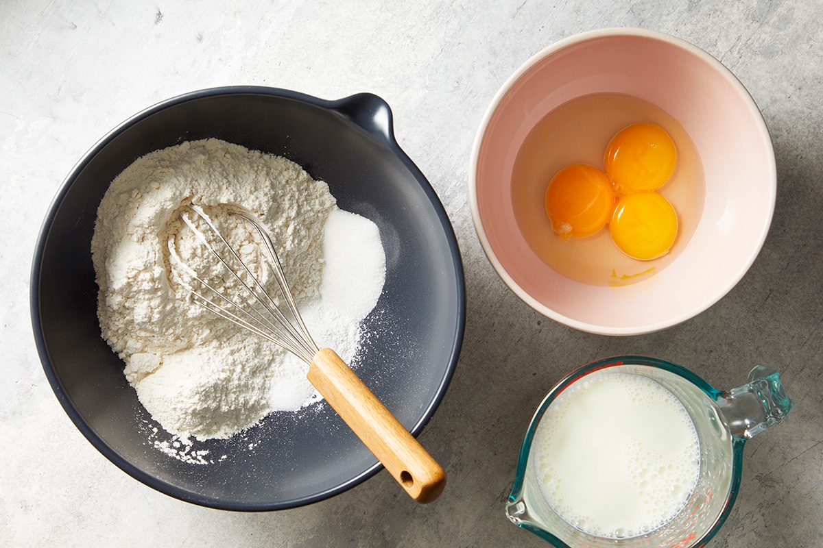 A whisk rests in a black bowl with flour and sugar. Next to it is a pink bowl holding three egg yolks and a glass measuring cup filled with milk, all on a light gray surface.