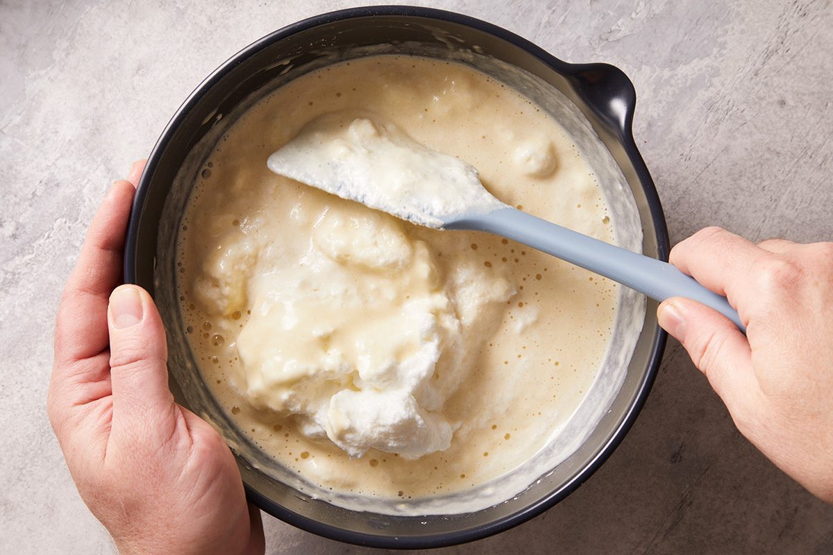 A person holds a mixing bowl and uses a spatula to fold whipped egg whites into a light, creamy batter on a gray surface.