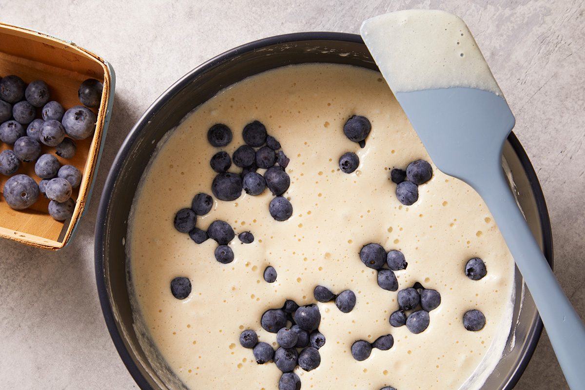 A mixing bowl filled with pancake batter and fresh blueberries, with a spatula resting on the edge. 