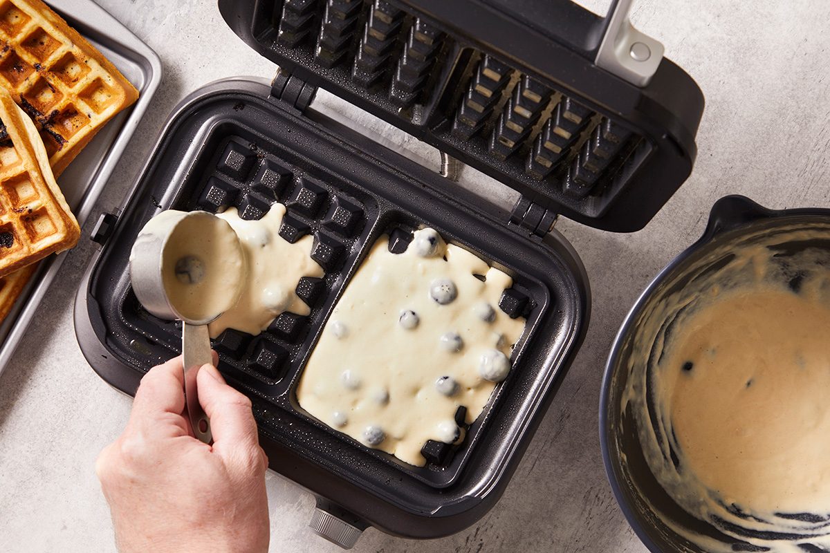 A hand pours blueberry waffle batter from a measuring cup onto a heated waffle maker; baked waffles and a bowl of batter are nearby on the counter.
