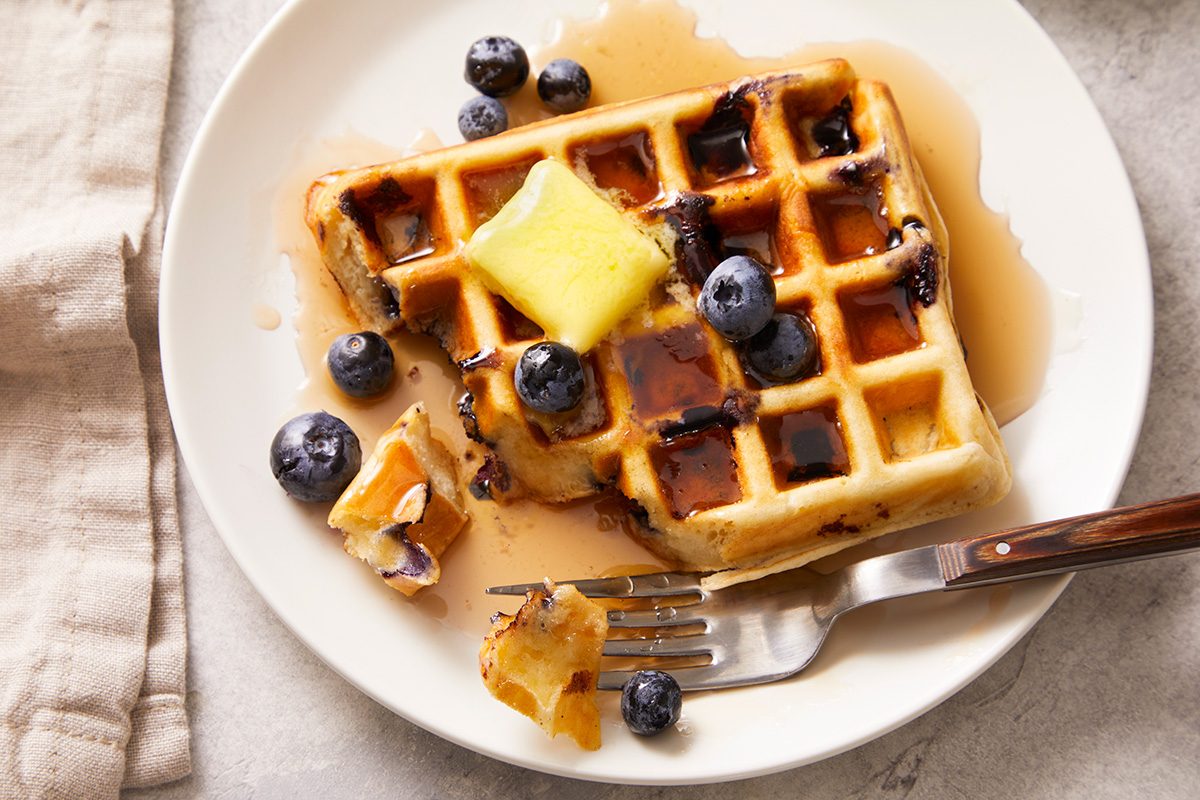 A partially eaten blueberry waffle topped with butter, syrup, and fresh blueberries sits on a white plate with a fork and beige napkin nearby.