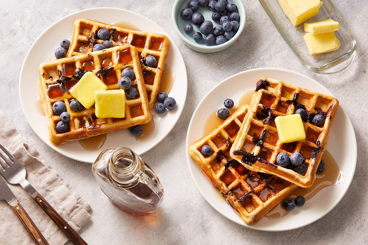 Two plates of waffles topped with butter, blueberries, and syrup sit on a table next to a glass syrup bottle, extra blueberries in a small bowl, and a dish of butter. Forks and a napkin are beside the plates.