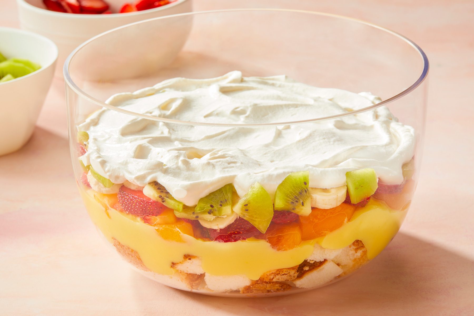 horizontal shot of fruit salad with a layer of cream on top presented in a large, clear bowl; the background shows additional bowls containing more fruits