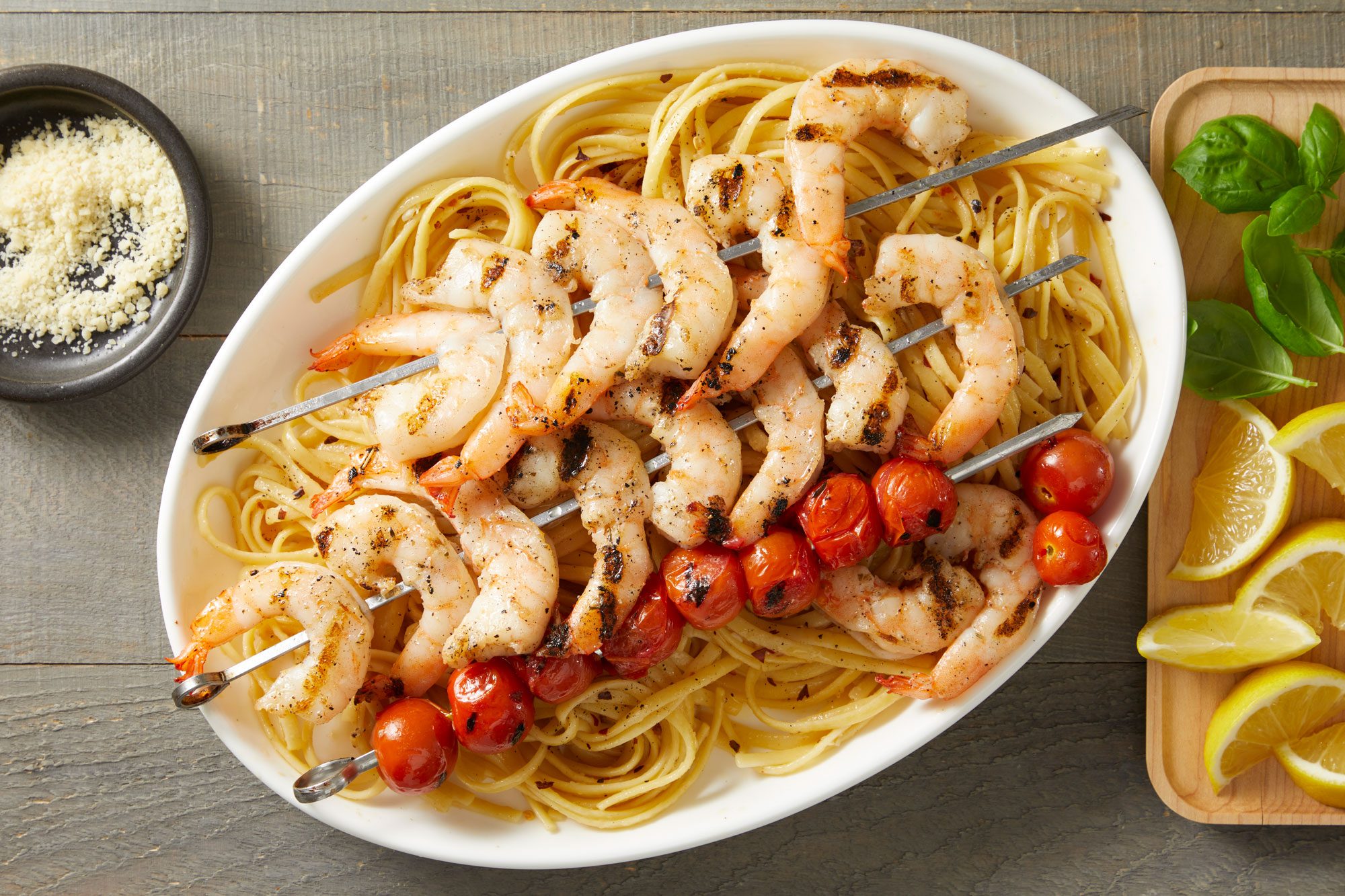 overhead shot of Grilled Shrimp & Tomatoes with Linguine in a white oval platter filled with a generous serving of linguine topped with grilled shrimp skewers and roasted cherry tomatoes; on the side, there is a small black dish of grated cheese, fresh basil leaves, and lemon wedges arranged neatly on a wooden cutting board