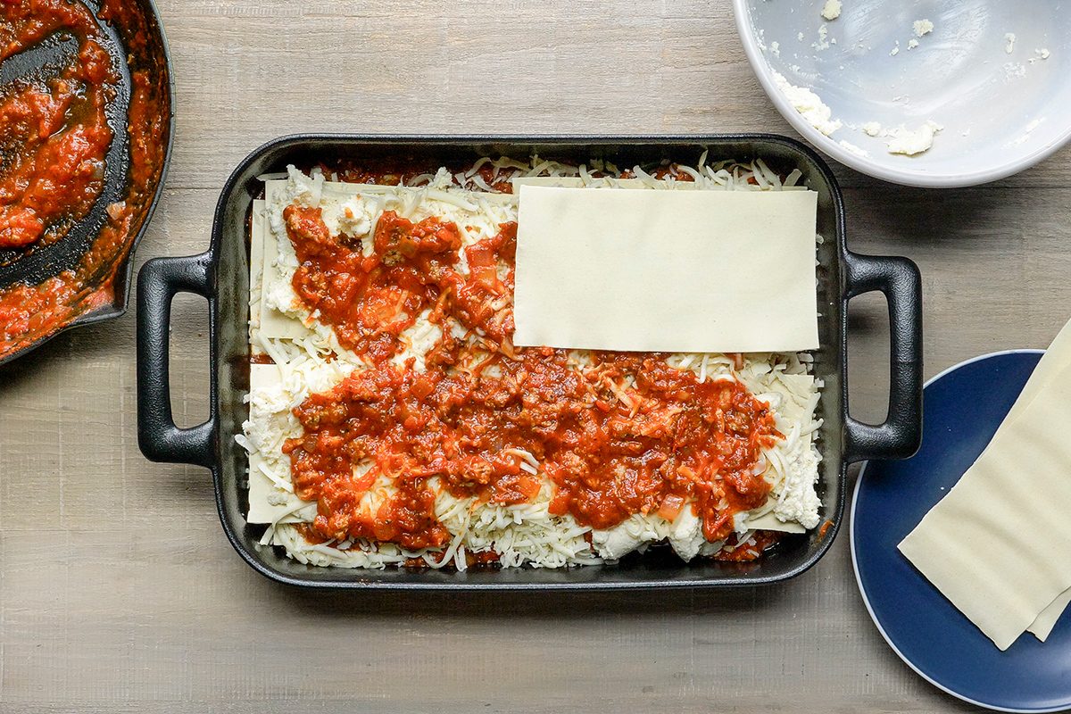 A lasagna being assembled in a black baking dish on a wooden surface. Layers of cheese and meat sauce are visible. Nearby, there's a round, empty pan with sauce remnants and a white plate with unused lasagna sheets.
