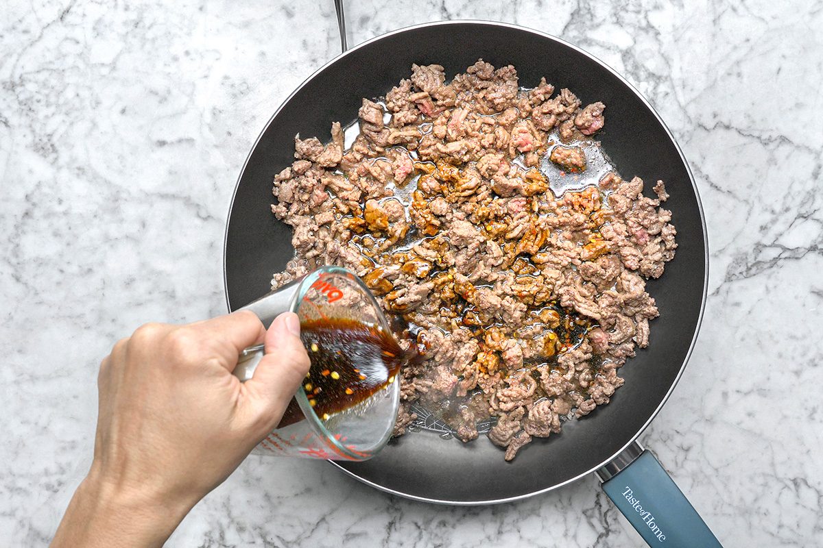 A person pours a dark sauce from a measuring cup onto cooked ground beef in a nonstick skillet. The skillet is on a marble countertop.
