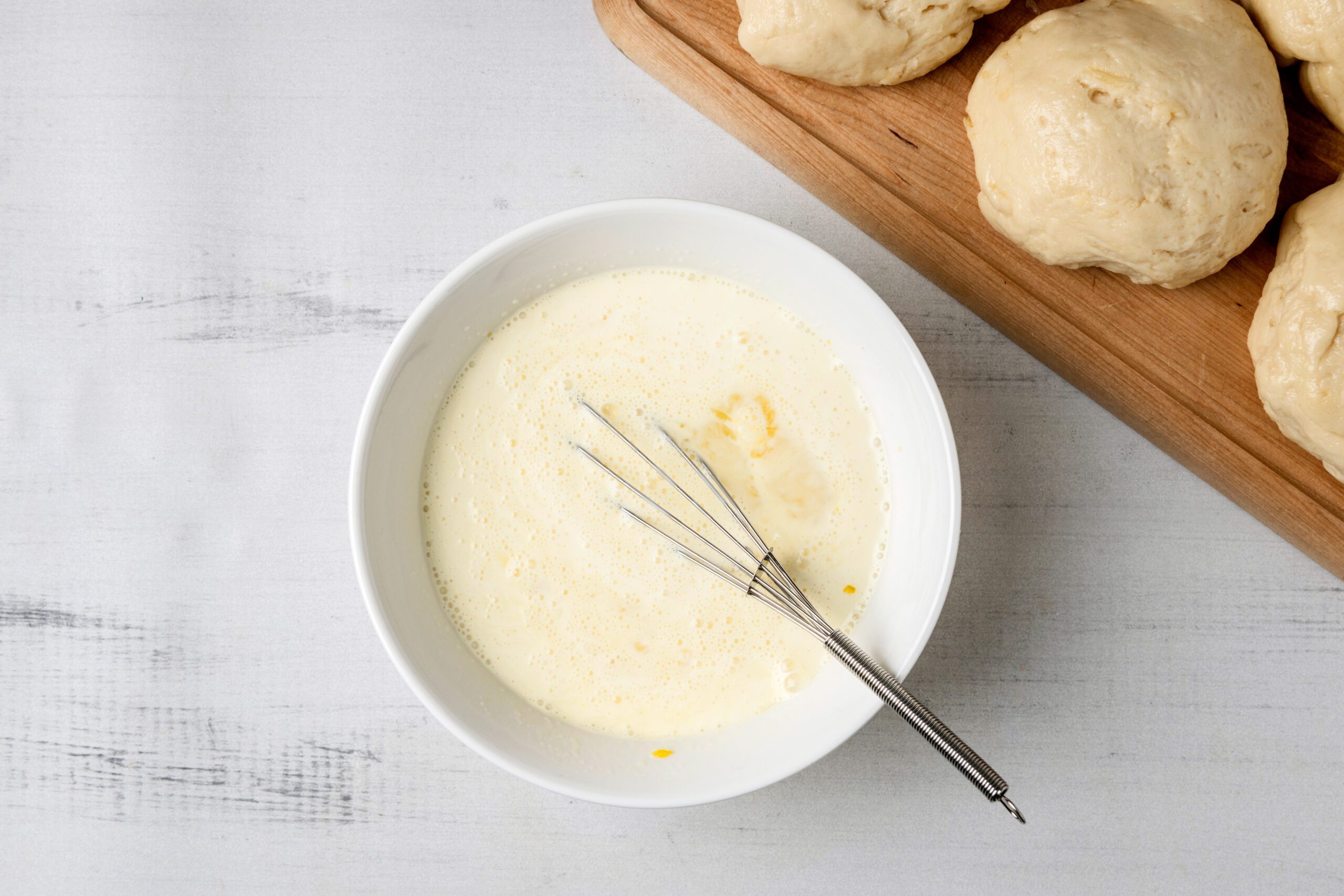 A white bowl containing a creamy batter with a small whisk rests on a light surface. Next to it, several pieces of unbaked dough are placed on a wooden board.