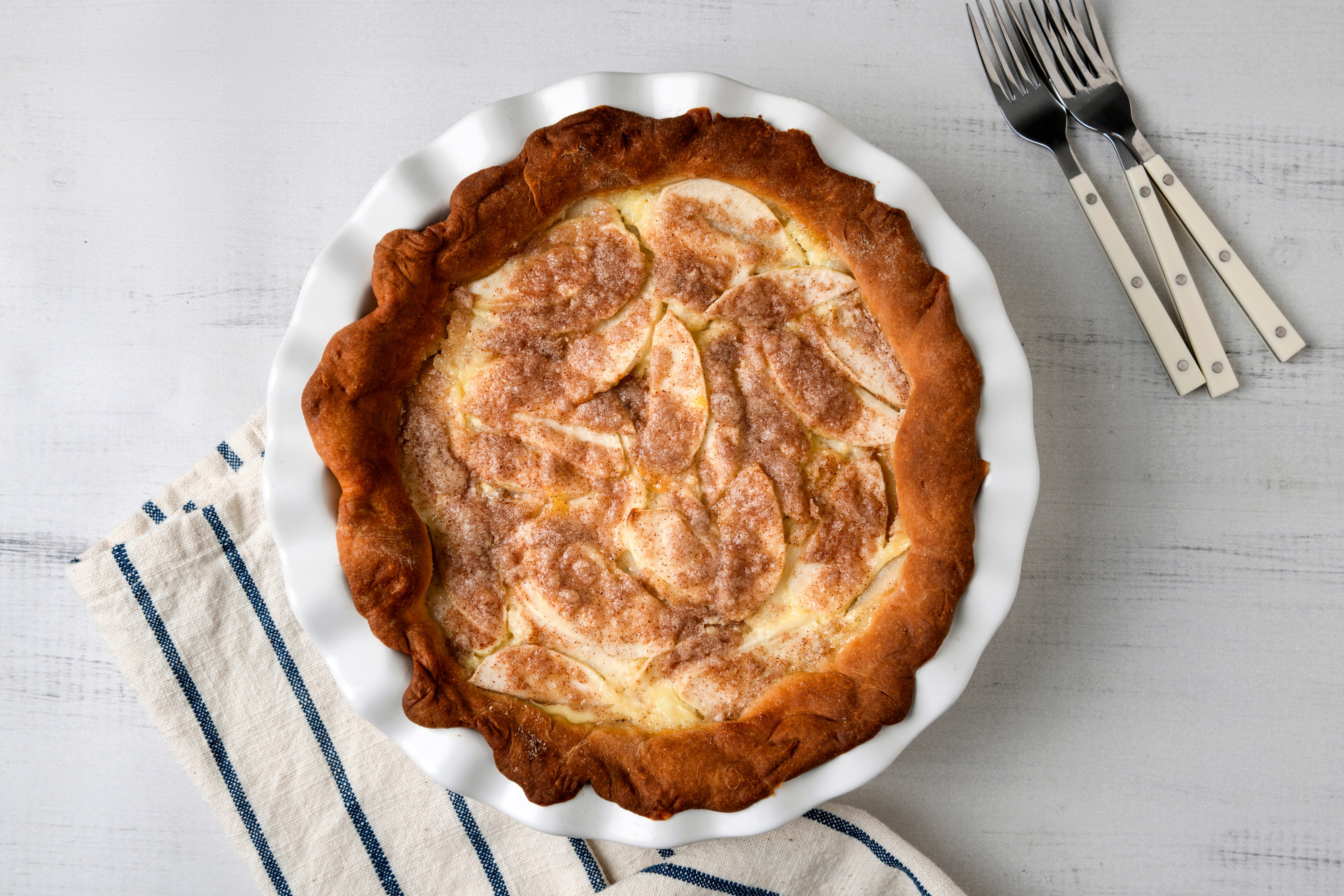 A freshly baked apple pie with a golden crust sits in a white fluted pie dish. The pie is topped with cinnamon-sprinkled apple slices. It rests on a white surface with a striped cloth. Three forks lie nearby.