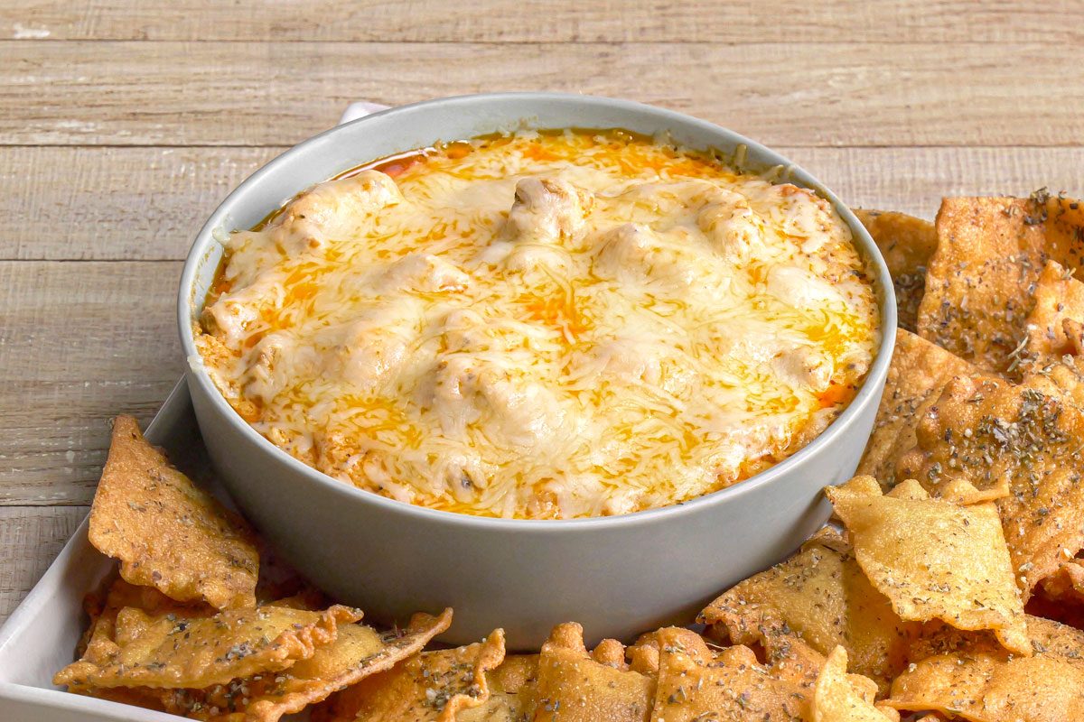 3/4th shot of Lasagna Dip served in a circular bowl placed on a rectangular platter; the dip is topped with melted cheese, surrounding the dip are crispy, golden brown ravioli chips, the background is wooden