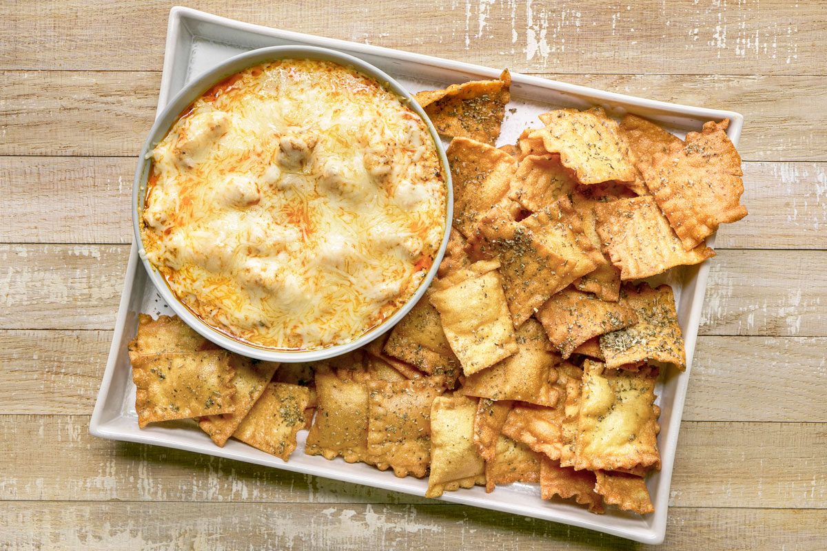 overhead shot of Lasagna Dip served in a circular bowl placed on a rectangular platter; the dip is topped with melted cheese, surrounding the dip are crispy, golden brown ravioli chips, the background is wooden