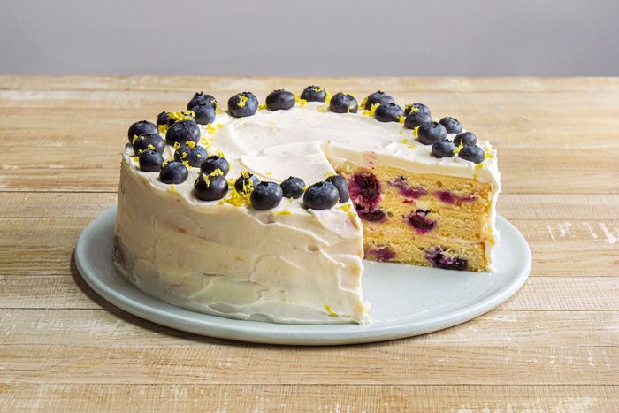 3/4th shot of lemon blueberry cake placed on a light colored round plate, the cake is adorned with a layer of frosting and decorated with fresh blueberries and some yellow zest on top, a slice has been removed from the cake, the background consists of a wooden surface