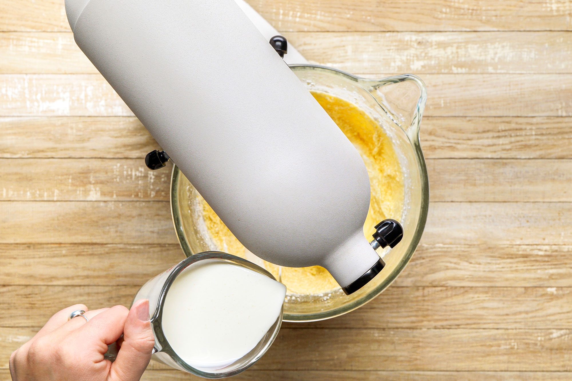 overhead shot of a hand is holding a measuring cup filled with a creamy liquid, ready to be poured into a mixing bowl below; the bowl contains a yellow mixture, likely a batter or dough, currently being blended by a stand mixer positioned above it
