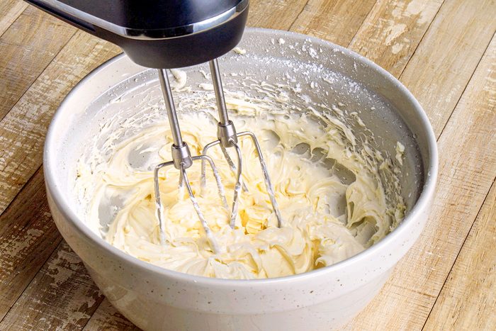 3/4th shot of a mixing bowl containing creamy batter; an electric mixer with metallic beaters is inserted into the mixture, a wooden surface background