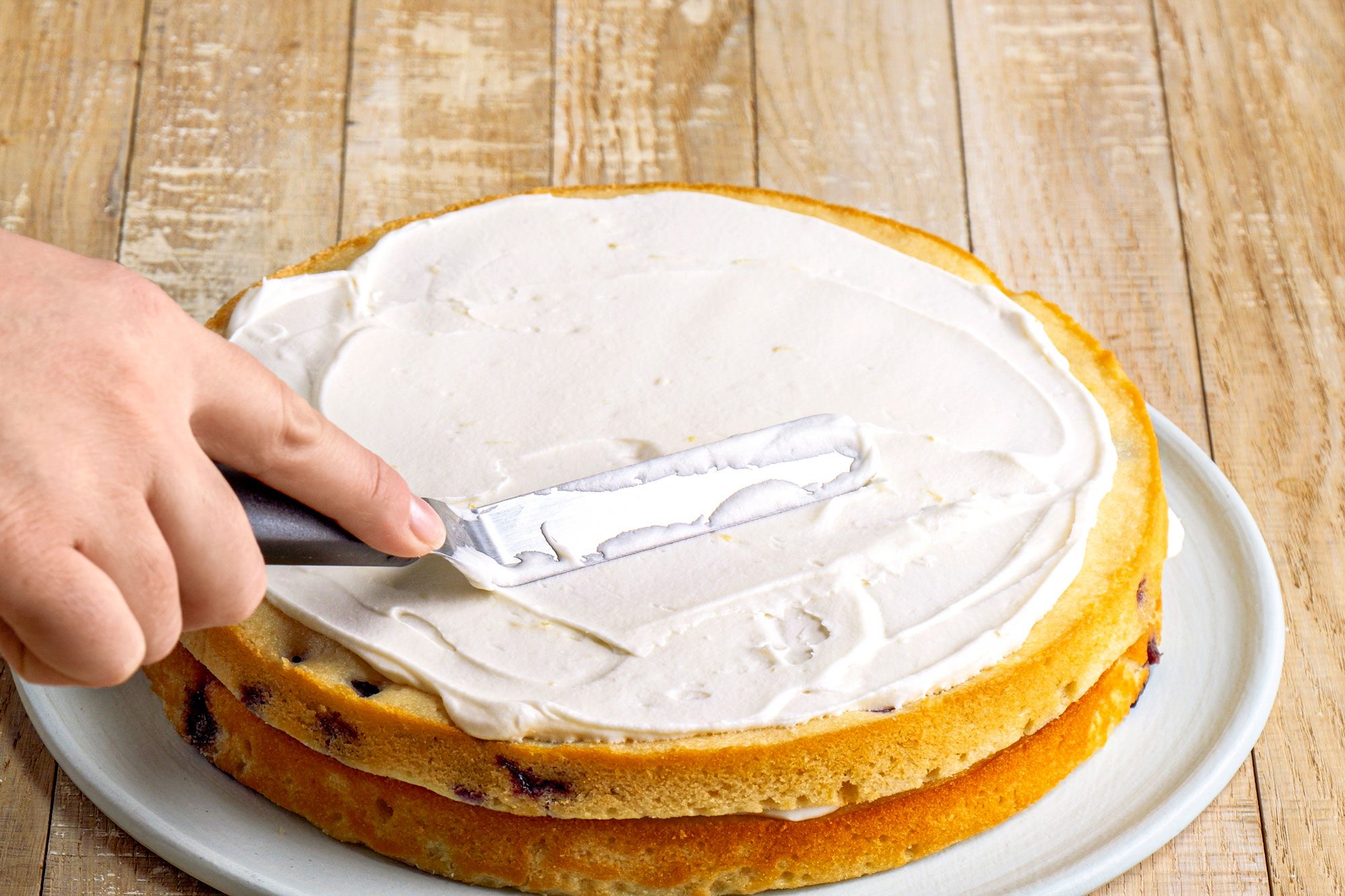 3/4th shot of a hand is seen spreading frosting onto a two layer cake using a spatula; the cake appears to be on a circular white plate placed on a wooden surface;