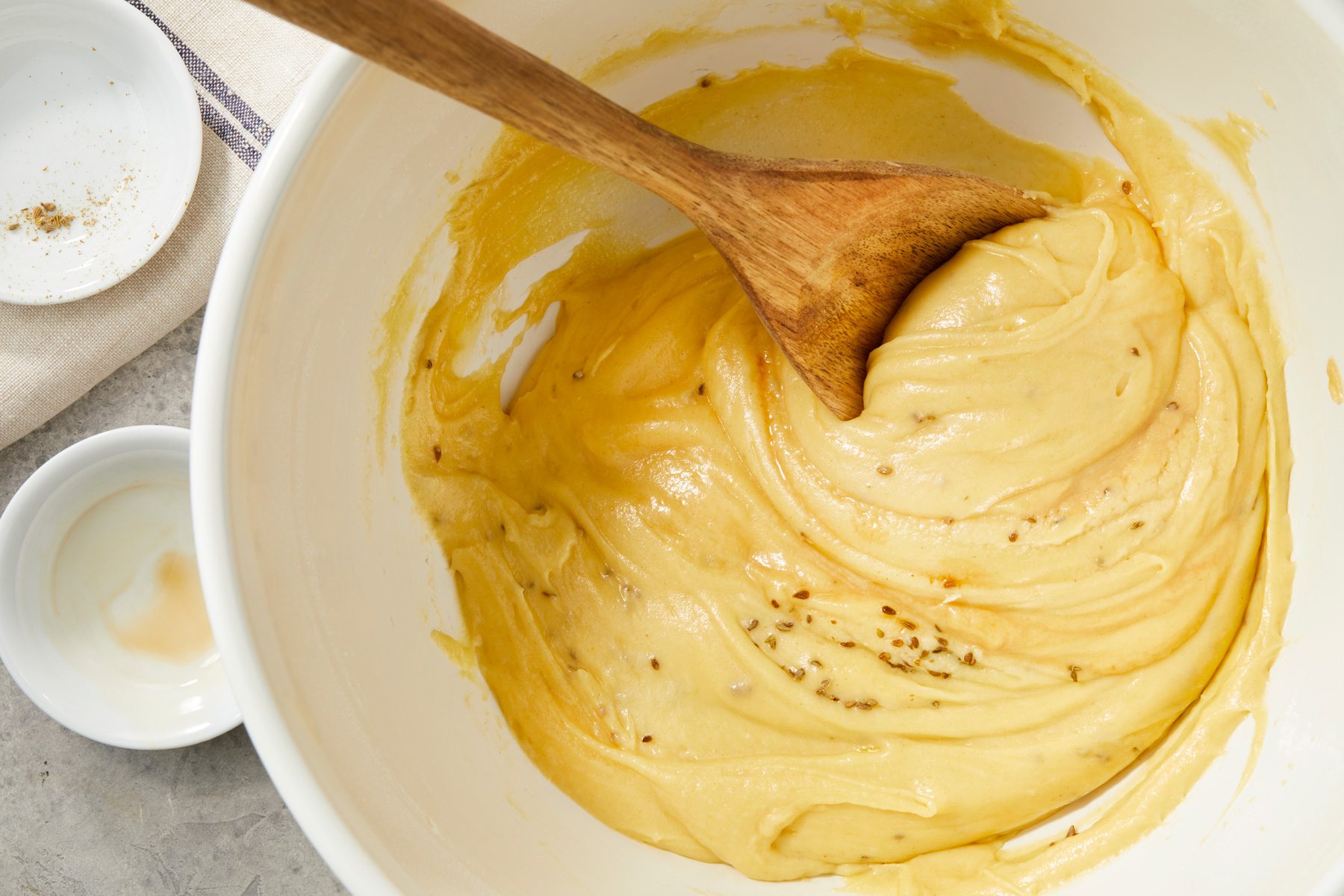 overhead shot of a mixing bowl filled with a creamy yellow batter, a wooden spoon is partially submerged in the mixture, in the background, there is a small white dish containing a translucent liquid, alongside a beige cloth with a subtle striped pattern