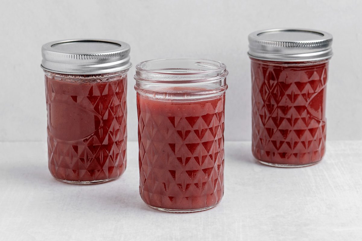 Three glass jars with a diamond pattern are filled with reddish-pink liquid. Two jars have metal lids, while the jar in the center is open. The jars are placed on a light-colored surface.
