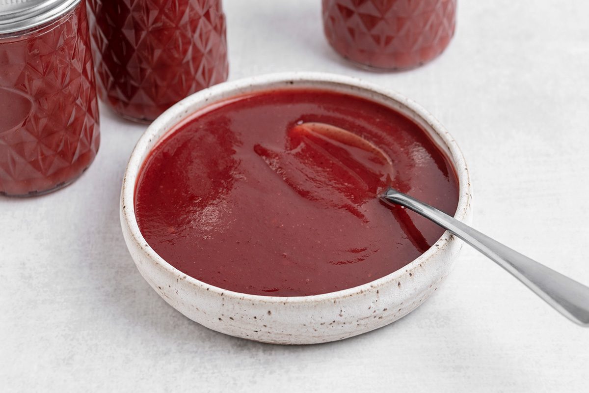 A white bowl filled with smooth, red ketchup, with a spoon resting inside. In the background, there are jars containing more ketchup.