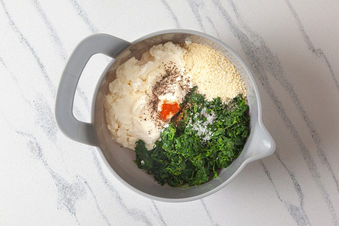 A gray bowl on a marbled surface contains ingredients for a dip: white creamy mixture, chopped green spinach, breadcrumbs, and spices, including salt and pepper.