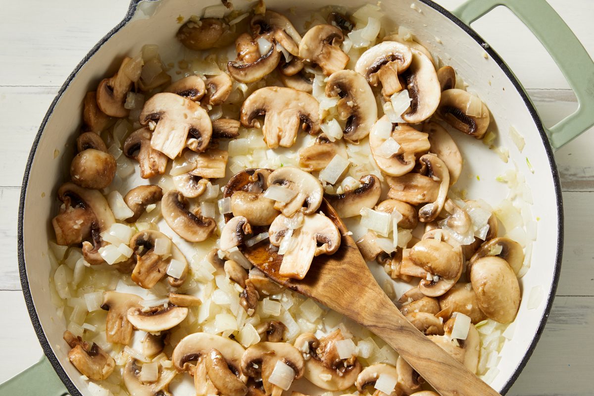 Overhead shot of cook pasta according to package directions; Meanwhile saute mushrooms and onion in oil in a large skillet until tender; add garlic; cook 1 minute longer; wooden spoon; white wooden surface;