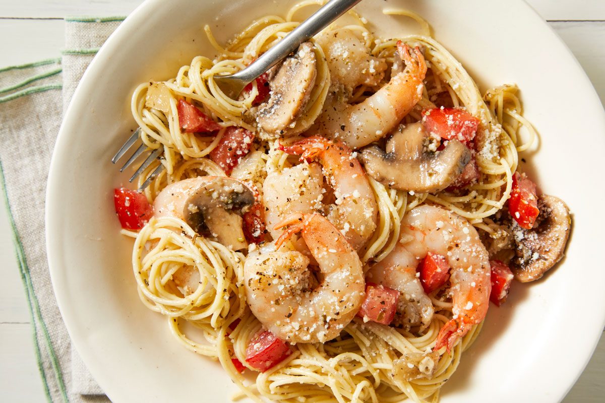 Close-up shot of Garlic Shrimp & Mushroom Pasta; served in a bowl with fork; sprinkle cheese on top; napkin; white wooden surface;