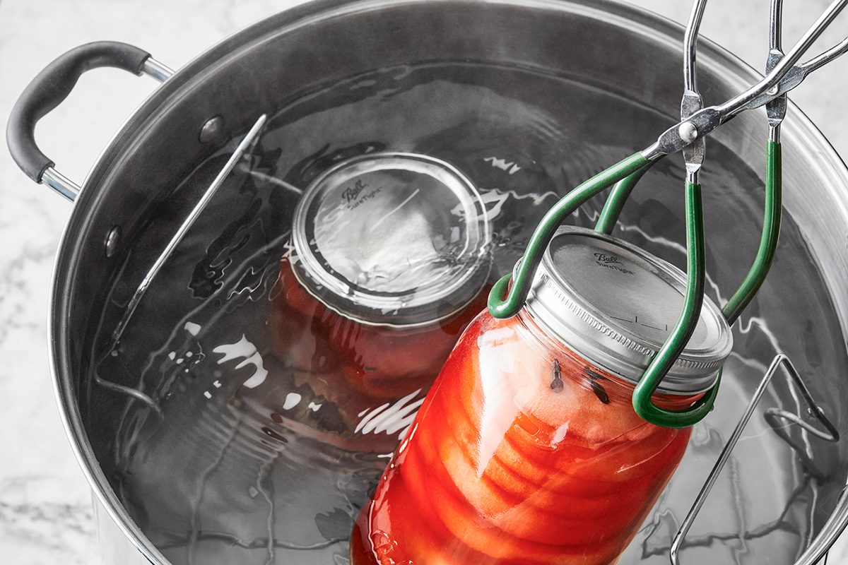 A glass jar filled with sliced vegetables is being lifted from a large pot of hot water with jar tongs during a canning process. Another jar remains submerged in the pot.