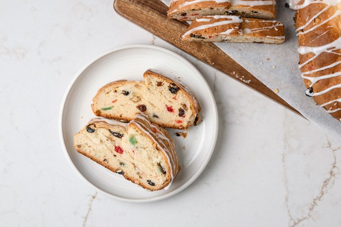 overhead shot of baked bread slices on a white plate; adorned with colorful bits of dried fruit and nuts; a thin drizzle of icing runs decoratively over the top of the slices, behind the plate, additional slices rest on a wooden board, while the background features a clean marble surface