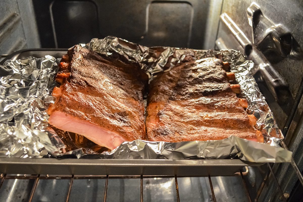 A rack of ribs covered in seasoning is baking on a foil-lined tray inside an oven. The oven door is open, showing the ribs roasting on the middle rack.