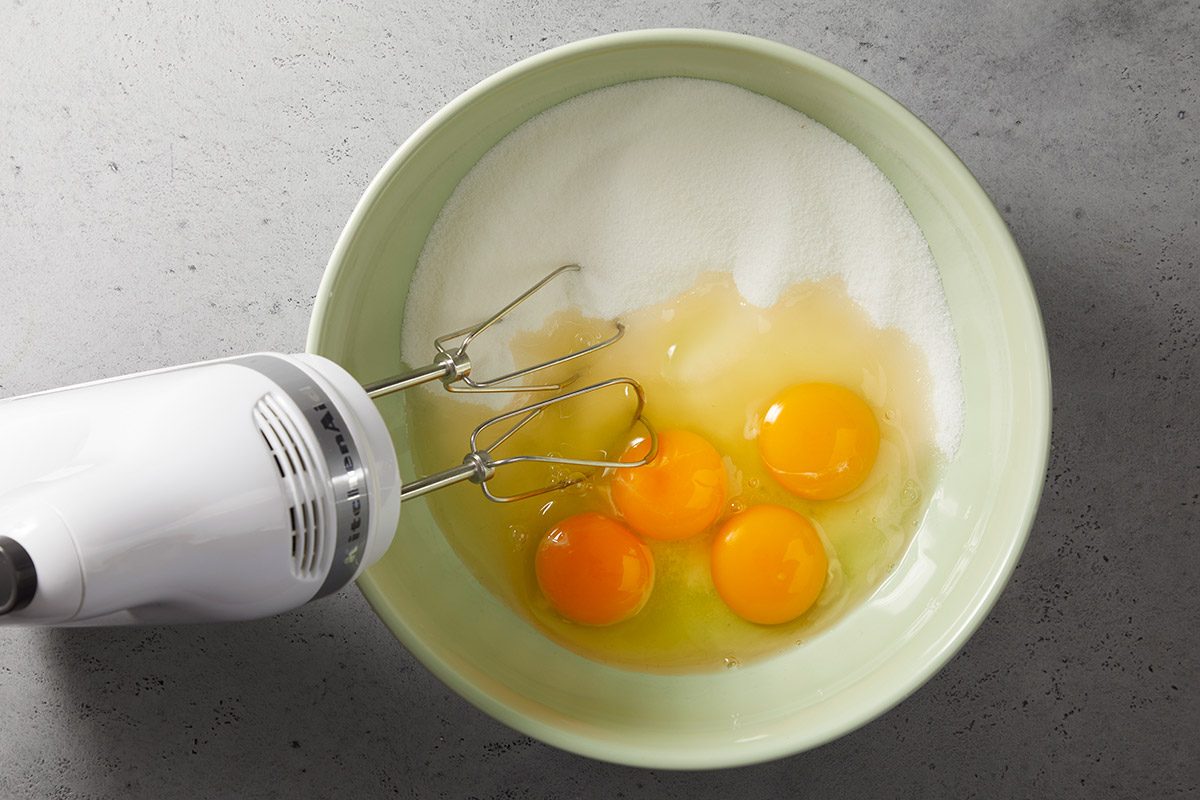 A hand mixer with beaters is positioned in a bowl containing three raw eggs and sugar, ready to be mixed. The bowl sits on a gray countertop.