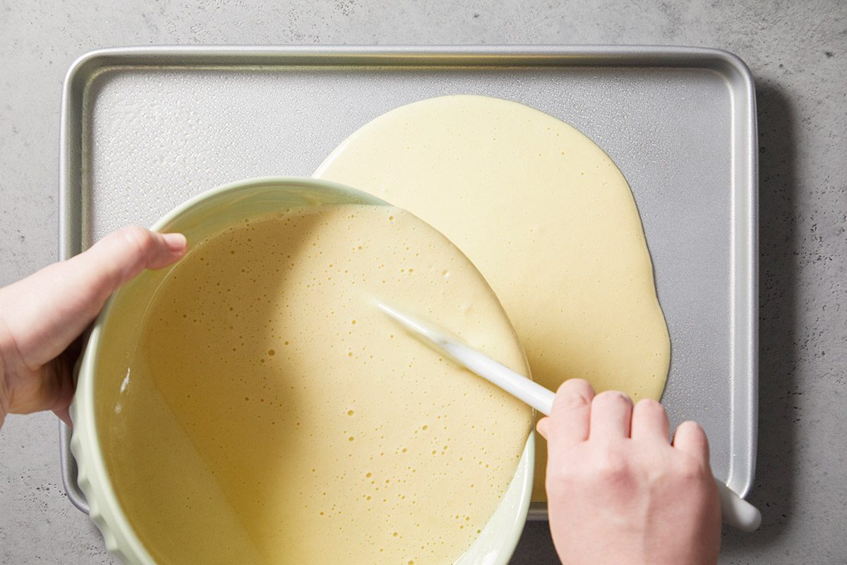 A person pours a bowl of smooth, pale batter onto a baking sheet, spreading it with a spatula on a gray countertop.