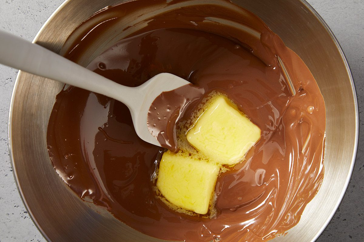 A metal bowl with melted chocolate and two cubes of butter being stirred with a white spatula.