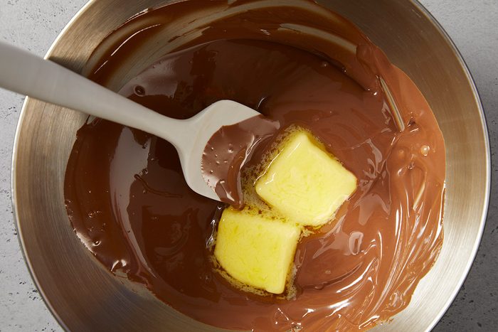 A metal bowl with melted chocolate and two cubes of butter being stirred with a white spatula.