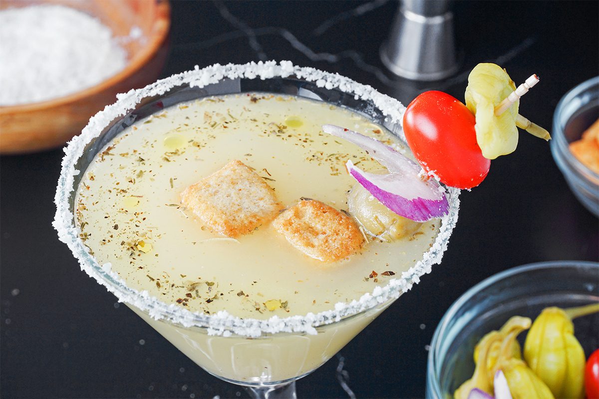 A cocktail glass filled with a light yellow drink, garnished with a salted rim. The drink is topped with croutons, herbs, and garnished with red onion, a cherry tomato, and a pepper on a skewer. Other ingredients are visible in the background.