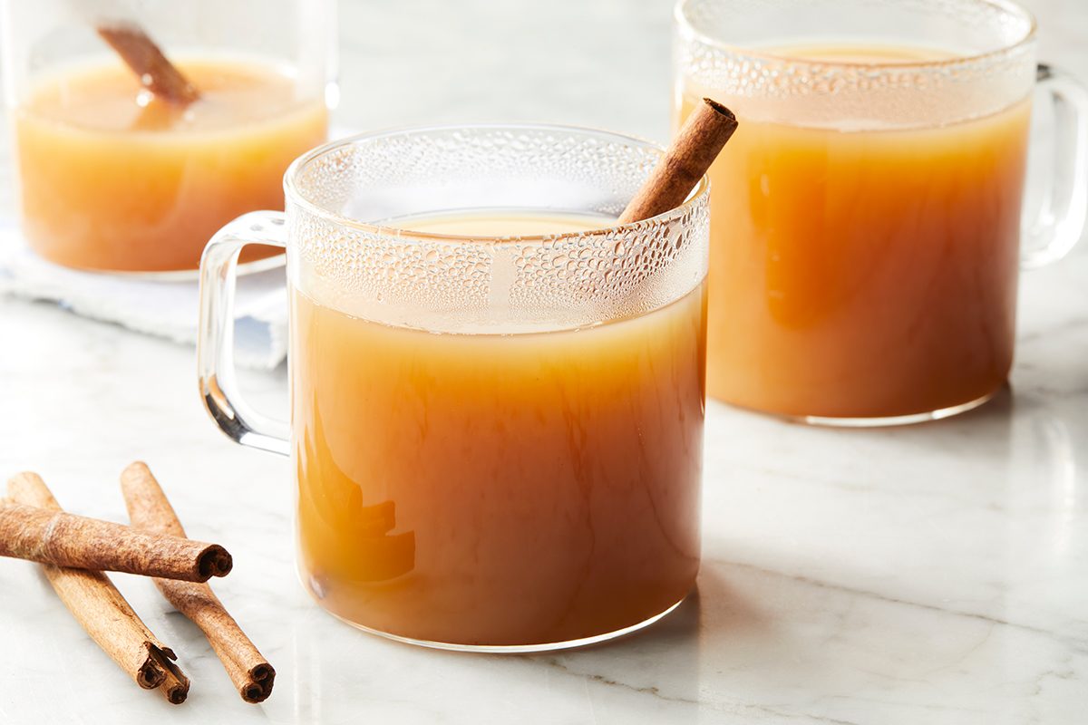 Three clear glass mugs filled with a warm, light brown beverage, each garnished with a cinnamon stick. Two cinnamon sticks lay on a white marble surface nearby.