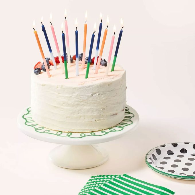 White cake with multicolored candles and sprinkles on a Kate Spade ceramic cake stand on a tan background