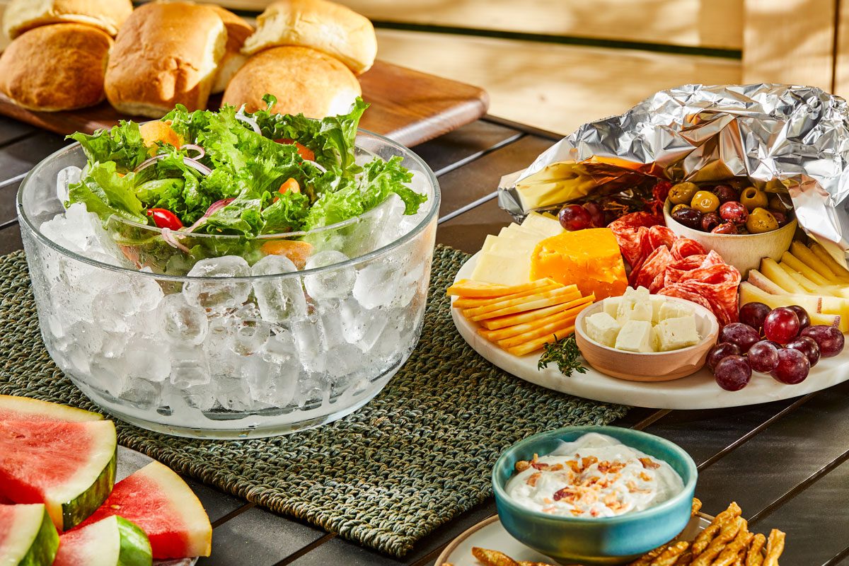 a beautifully arranged outdoor dining spread, featuring a crisp salad in a large glass bowl sitting on a bed of ice, colorful greens and vegetables are visible throughout; nearby, a platter is filled with an assortment of cheeses, meats, olives, and grapes, alongside slices of watermelon on a woven mat; fresh bread rolls are positioned in the background on a wooden board, while a small bowl of creamy dip garnished with bits of bacon
