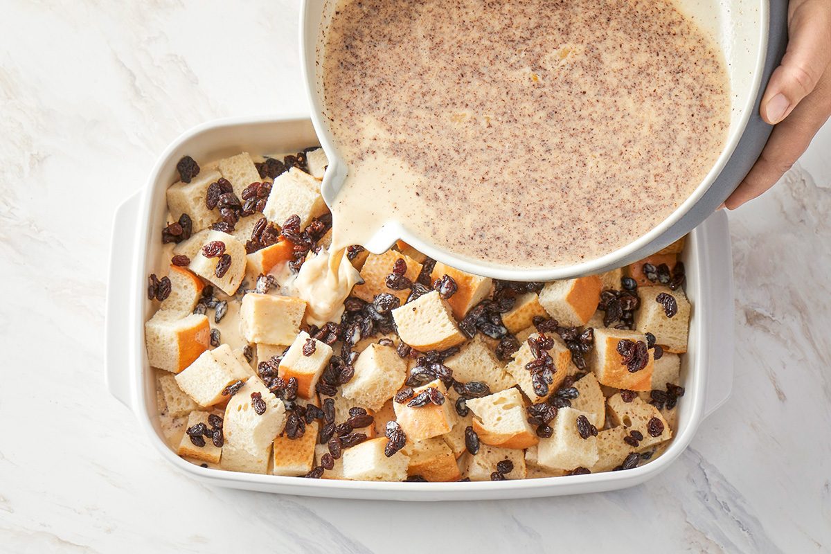 3/4th shot of A hand pouring a creamy egg mixture over cubed bread and raisins in a white baking dish, placed on a marble countertop