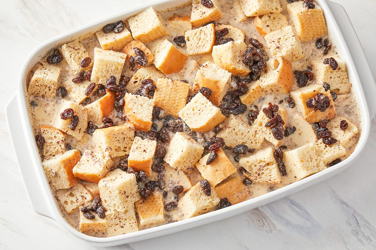 overhead shot of a white baking dish filled with cubed bread, raisins, and a creamy liquid mixture, ready to be baked, likely for bread pudding, The dish rests on a marble surface