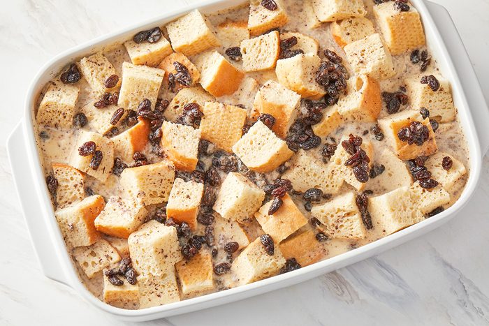 overhead shot of a white baking dish filled with cubed bread, raisins, and a creamy liquid mixture, ready to be baked, likely for bread pudding, The dish rests on a marble surface