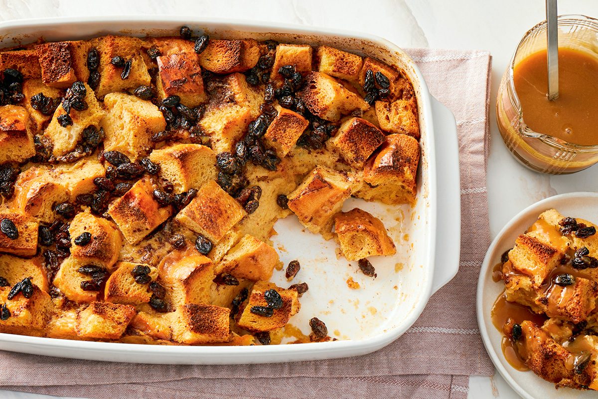 overhead shot of a white baking dish with read Pudding With Rum Sauce topped with raisins, partially served, A plate with a serving of bread pudding and a jar of caramel sauce sit nearby on a pink cloth