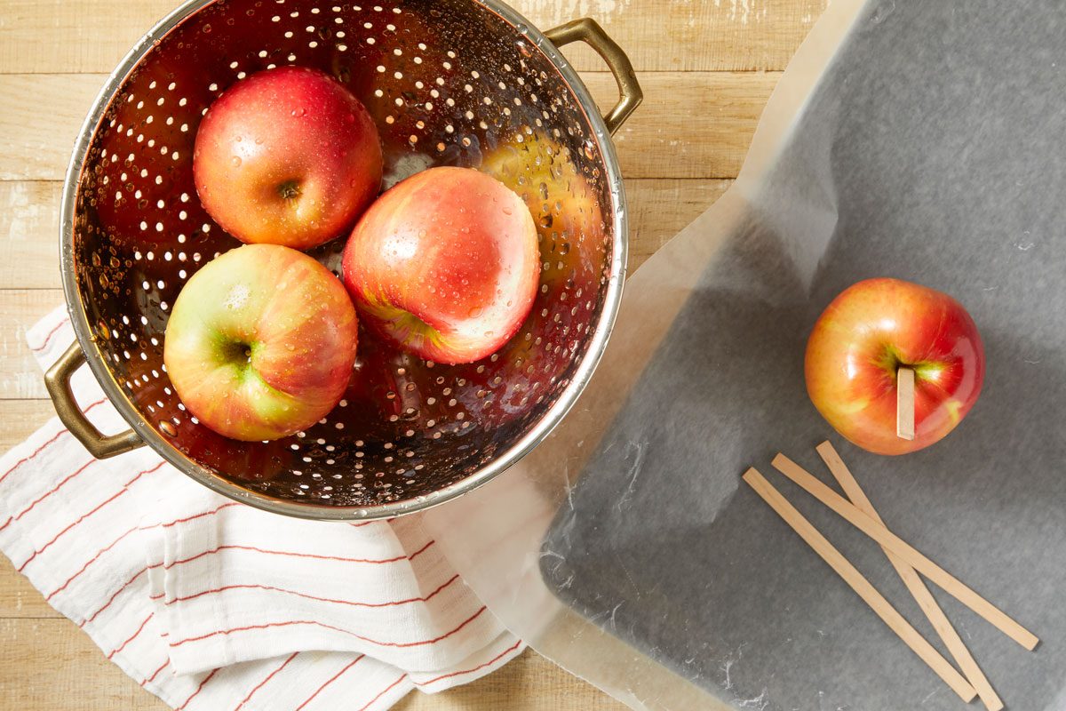 Overhead shot of wash and thoroughly dry apples; remove stems; Insert wooden pop sticks into apples; Place on a waxed paper-lined baking sheet; set aside; a napkin; all set on a light brown wooden surface;