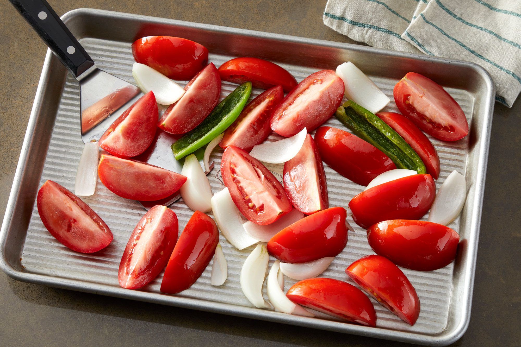 High angle shot; Preheat oven to 425 degree; Place tomatoes, onion and serrano on a rimmed baking sheet; Drizzle with oil; toss to coat; roast until vegetables are tender and lightly browned 15-20 minutes; Let slightly cool; flat spatula and a napkin on a light brown surface