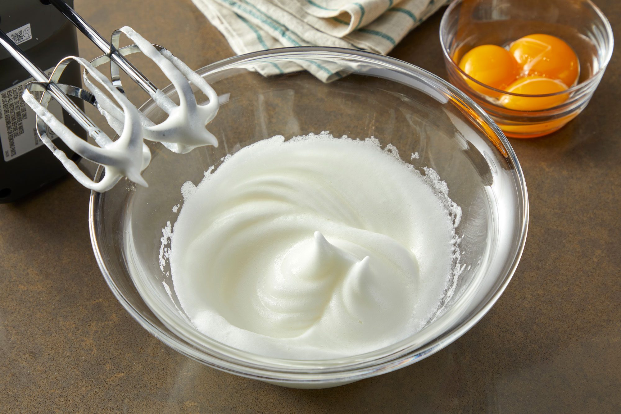 3/4 angle shot; Place egg whites in a large bowl; beat on high speed until stiff peaks form 3-4 minutes; On low speed beat in egg yolks until smooth; blender and a napkin is on light brown surface