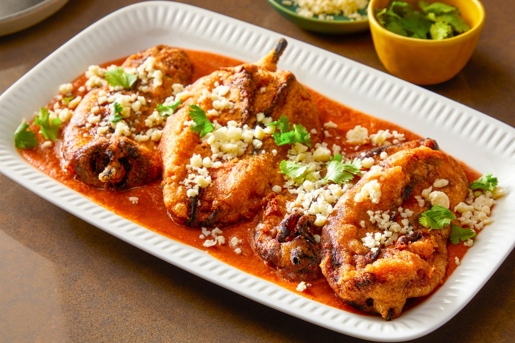 Table view shot of Chile Rellenos; To serve spoon red sauce in shallow bowls; top with fried peppers; Sprinkle with cilantro and Cotija cheese; Serve immediately; all arranged on a light brown surface