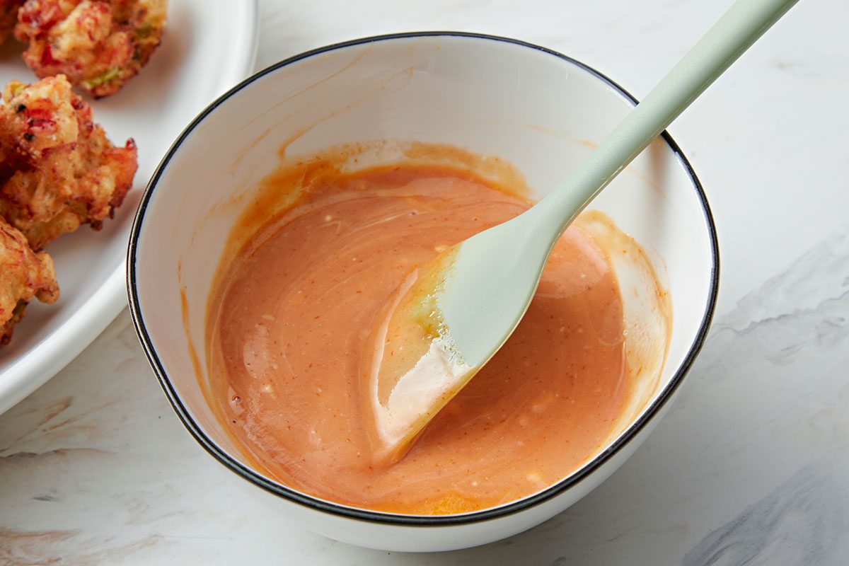 A white bowl filled with a creamy orange sauce, being stirred with a light green spatula, sits on a white surface next to a plate of battered food.