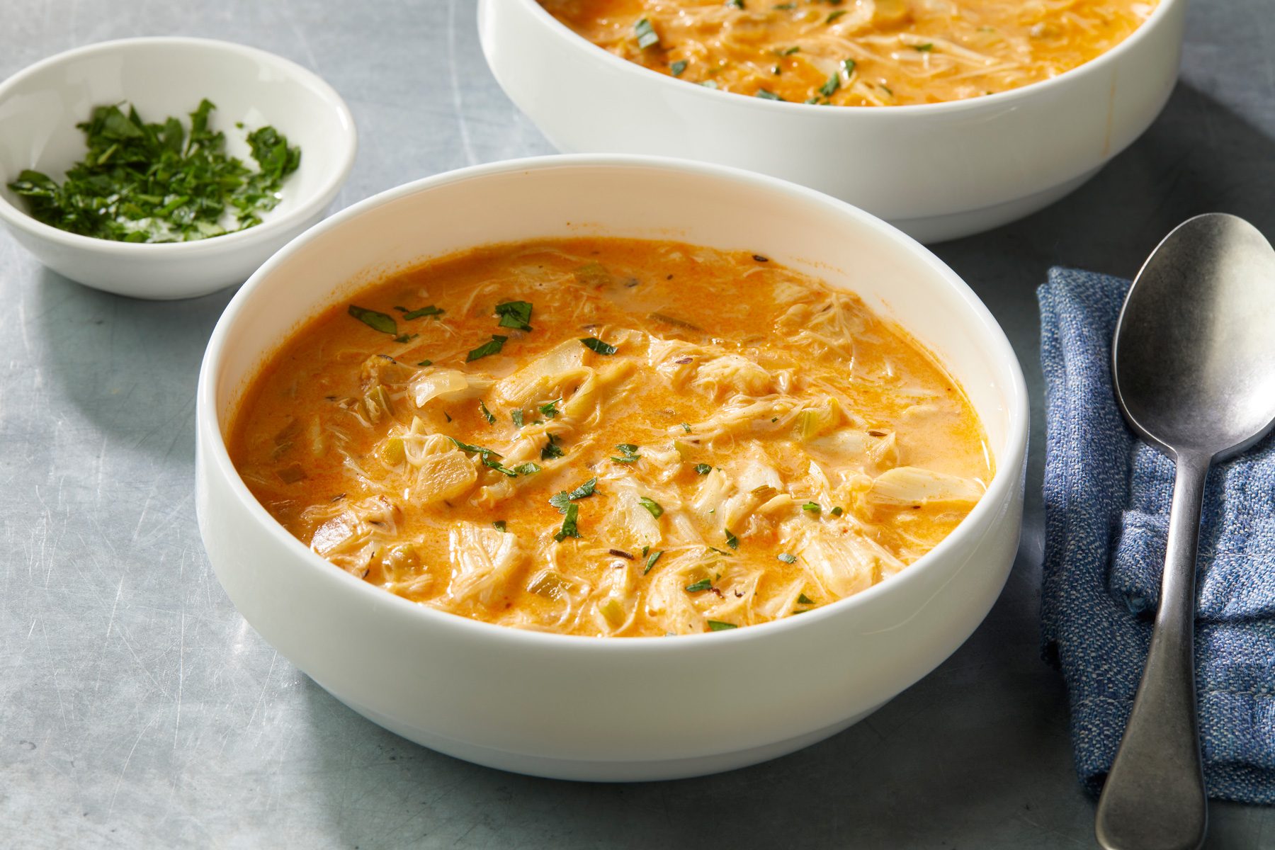 3/4 angle view shot of Homemade Crab Bisque; Served in two large bowls; Garnish with parsley; a spoon rests on a blue napkin; all set on a grey surface;