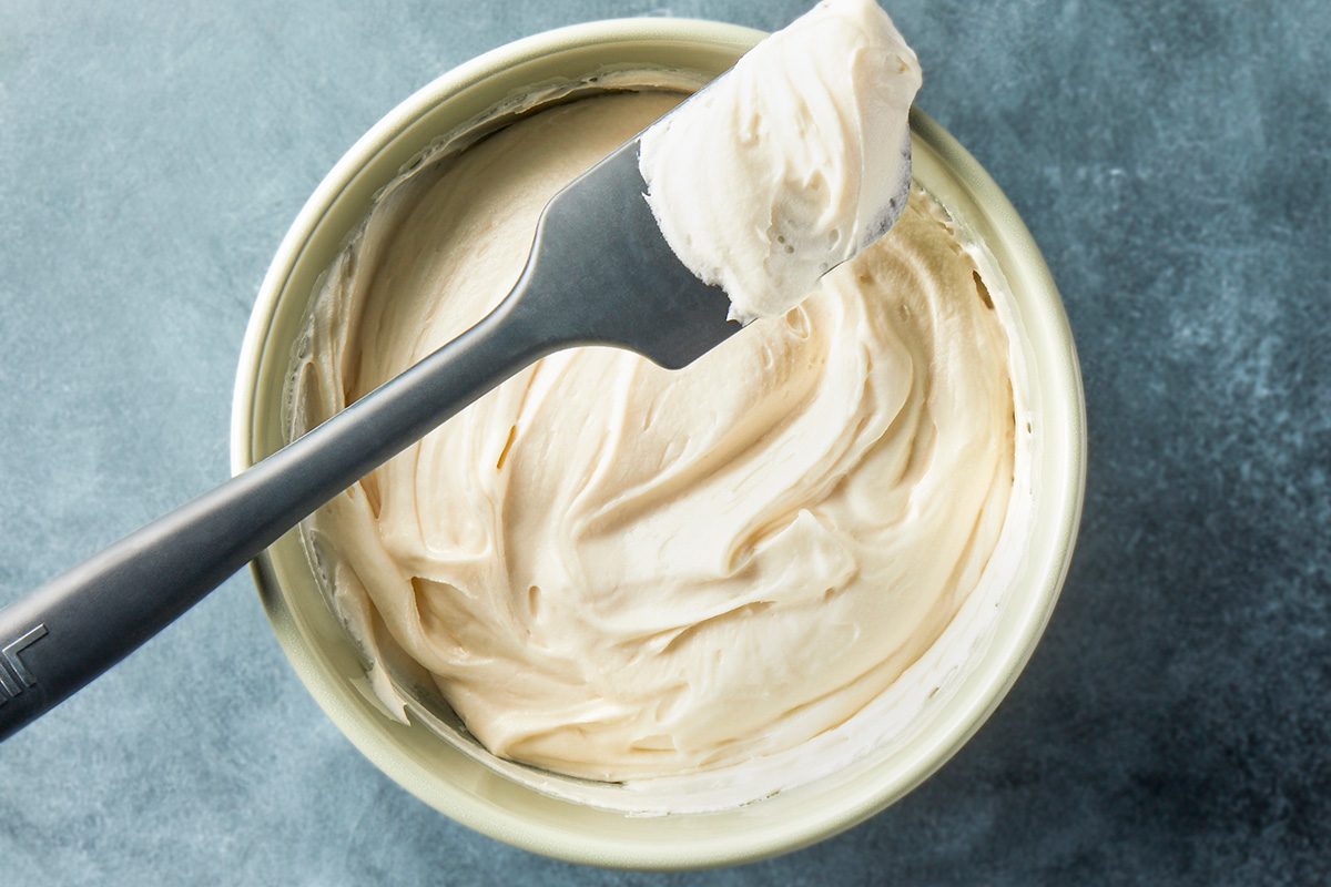 overhead shot of a bowl of creamy white frosting with a metal spatula resting on top, coated in frosting, on a gray textured surface